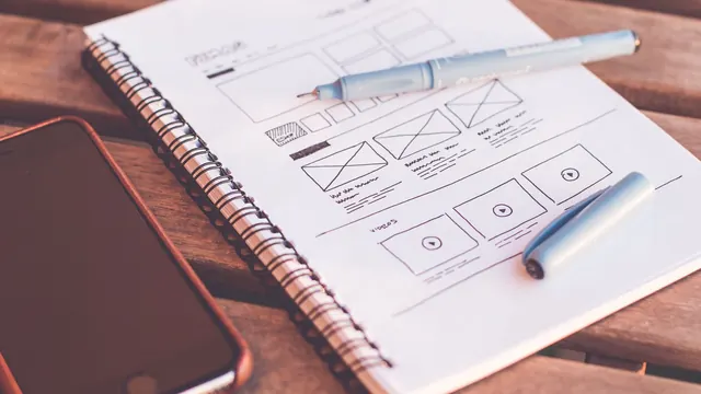 Notebook with website wireframe sketches beside a smartphone on a wooden desk